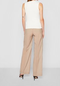 Mujer de pie de espaldas, vestida con una blusa sin mangas blanca, pantalones anchos beige, sandalias negras de tacón alto y un pequeño tatuaje en el brazo.