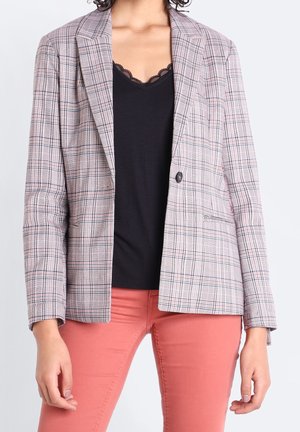 Kobieta ubrana w szarą kratowaną marynarkę na czarnym topie z koronkową lamówką oraz koralowych spodniach, stojąca na tle jasnego, jednolitego tła.