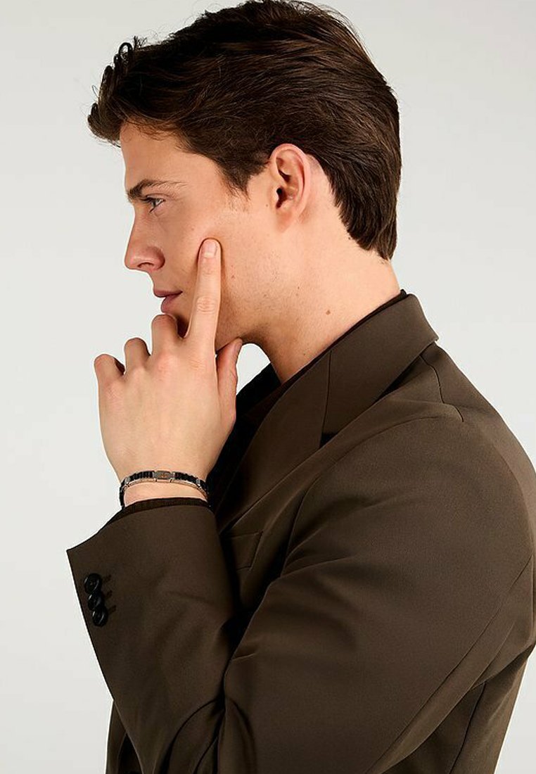 Mörkbrun blazer med slagkrage, bärs av en person med kort hår, som rör vid sitt ansikte med ett eftertänksamt uttryck och visar upp ett armband.