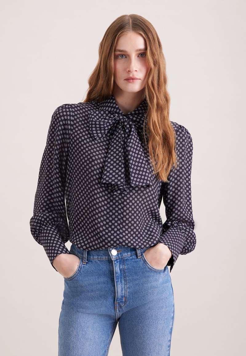 Femme aux longs cheveux ondulés portant un chemisier bleu marine à pois floraux blancs avec un col noué, associé à un jean taille haute bleu clair, mains dans les poches.