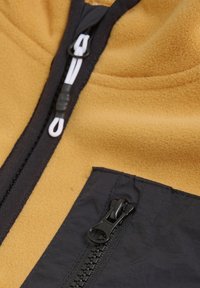 Gul fleece-stoff med en svart glidelåslomme og kontrasterende svart glidelås. Har hvit snor og en glatt tekstur.