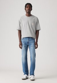 Graues Baumwoll-T-Shirt mit kurzen Ärmeln und einer Brusttasche; kombiniert mit hellblauen Jeans und weißen Sneakers mit schwarzen Akzenten.