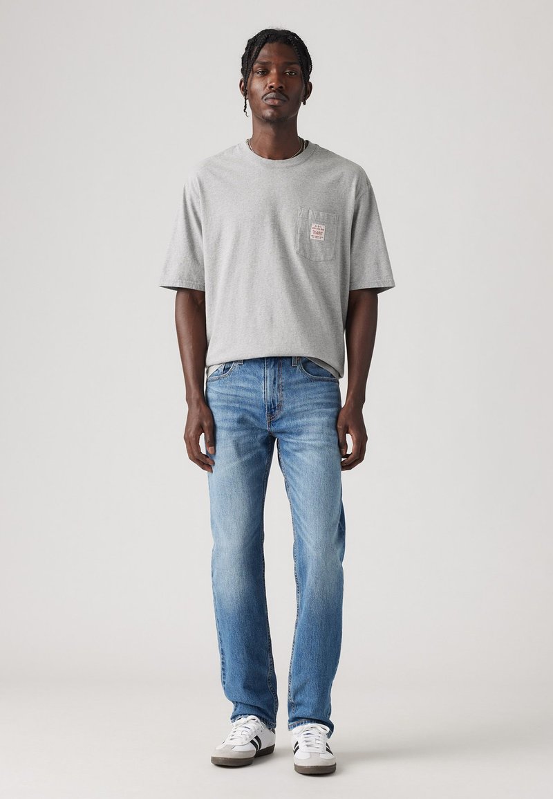 Graues Baumwoll-T-Shirt mit kurzen Ärmeln und einer Brusttasche; kombiniert mit hellblauen Jeans und weißen Sneakers mit schwarzen Akzenten.