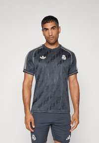 Jersey atlético de color gris oscuro con un patrón sutil, logo de Adidas, escudo del Real Madrid y rayas blancas contrastantes en las mangas.