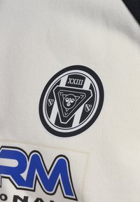 Μαύρο και άσπρο κυκλικό έμβλημα με λατινικούς αριθμούς XXIII, γεωμετρικά σχήματα και ένα στυλιζαρισμένο ζώο σε λευκό ύφασμα πάνω από μπλε κείμενο "RM".