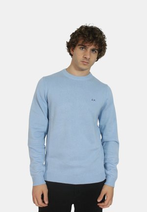 Maglione azzurro chiaro a girocollo in maglia con polsini e orlo ribattuti; piccolo logo sul petto; texture morbida. Indossato con pantaloni neri.