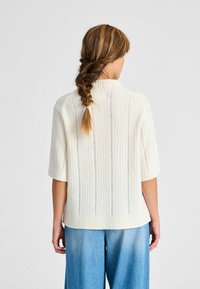 Krém színű bordázott kötött pulóver rövid ujjakkal, magas nyakkal és látható varrásokkal, világoskék farmernadrággal párosítva. Hátulról látható.