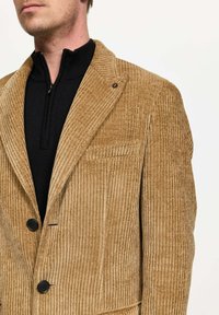 Blazer de pana en color marrón claro, con canalones verticales, solapas con muesca, dos botones negros y un bolsillo en el pecho. Llevado sobre una blusa negra.