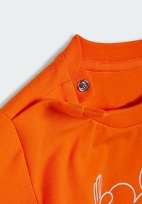 Pomerančové bavlnené tričko s výstrihom a patentným zapínaním, hladká textúra a čiastočne viditeľný biely dizajn na okraji.