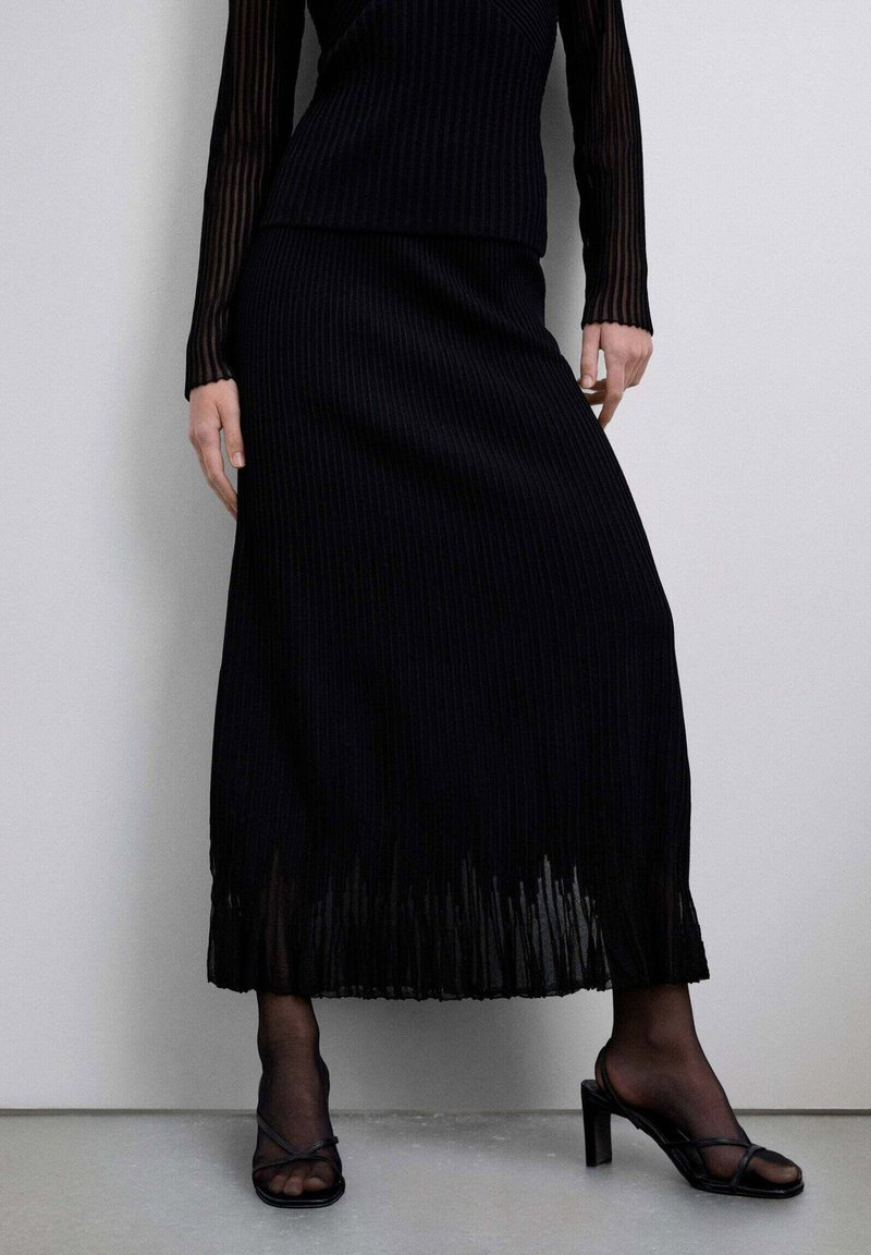 Conjunto de dos piezas de punto acanalado negro que incluye un top de manga larga y una falda larga hasta el suelo con dobladillo plisado, combinado con sandalias de tacón con tiras.