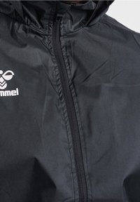 Giacca leggera nera con chiusura a zip, dotata di cappuccio e logo sul lato sinistro. Texture liscia e senza motivi visibili.