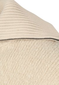 Close-up van beige gebreide stof met zichtbaar geribd patroon en een zwarte ritssluiting langs de naad.