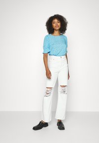 Blusa floral azul clara com mangas bufantes, combinada com calças de ganga brancas de perna larga com grandes rasgos nos joelhos, e sapatos pretos de slip-on.