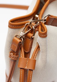 Beige väska med texturerad tyg, bruna läderdetaljer, gyllene hårdvara och en detaljerad stängningsmekanism. Har sydda läderremmar.