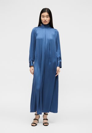 Naine seisab, kandes pikka, sinist satiinist kõrge kaelusega kleiti pikkade varrukatega ja musti avatud varbaga kõrgete kontsadega sandaale ühevärvilise tausta ees.