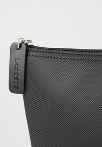 Gros plan sur une pochette zippée noire texturée avec un tirette en caoutchouc emboutie du lettrage "LACOSTE".