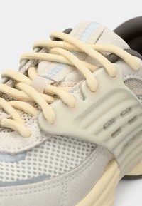 Gros plan d'une basket beige en tissu mesh texturé, lacets crème et support latéral en plastique sur une semelle claire.