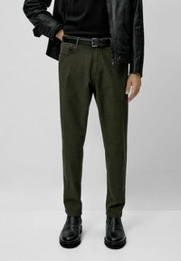 Tummanvihreät puuvilla-chinonhousut, kapealahkeiset, sivutaskuilla ja vyöllä. Yhdistetty mustaan nahkatakkiin ja kiiltäviin mustiin nyörisaappaisiin.