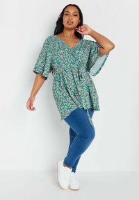 Top floreale avvolgente con scollatura a V, maniche corte a campana e vita con lacci, abbinato a jeans skinny blu e sneakers bianche. Il motivo presenta fiori multicolori.