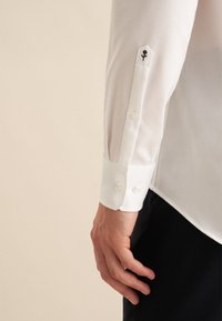 Witte blouse met lange mouwen van textuurstof, voorzien van een spitse manchet, twee knopen en een zwart geborduurd bloemenmotief op de mouw.