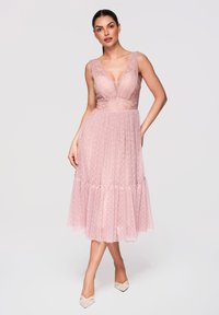 Femme portant une robe midi rose en dentelle et tulle sans manches à pois, posant avec une main sur la hanche et des talons blancs pointus.