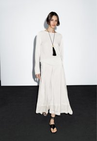 Lichtbeige shirt met lange mouwen en een vloeiende rok met een zoomlijn. Een zwarte ketting met kwastje zorgt voor contrast. Model staat tegen een effen achtergrond.