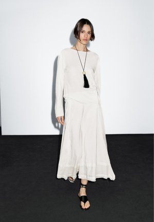 Ljusbeige långärmad skjorta och flödande kjol med fåll. Ett svart tofs-halsband skapar kontrast. Modellen står framför en enkel bakgrund.