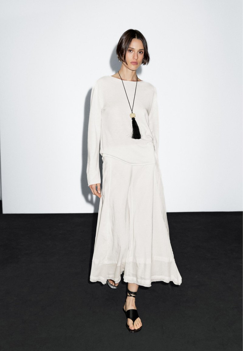 Hellbeige langärmeliges Hemd und fließender Rock mit Saum. Eine schwarze Quasten-Halskette sorgt für Kontrast. Das Model steht vor einem schlichten Hintergrund.