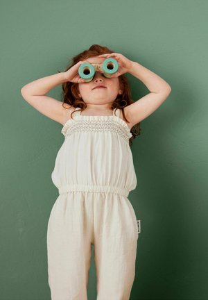 Salopette blanche sans manches avec détail froncé, taille élastique et jambes amples. Un enfant tient des jumelles de teinte verte devant un fond vert.