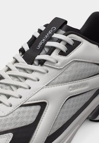 Άσπρο και γκρι αθλητικό παπούτσι Calvin Klein με λευκά κορδόνια, μαύρες λεπτομέρειες και λογότυπο Calvin Klein στη γλώσσα και στην πλαϊνή πλευρά.