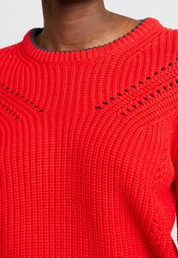 Rød strikket sweater med tekstureret rib, rund halsudskæring med kontrastfuld marineblå kant og indviklet hulmønster.