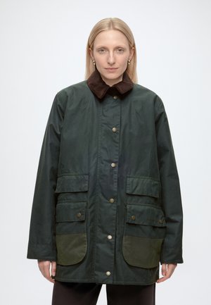 Frau mit glatten blonden Haaren trägt eine dunkelgrüne Jacke mit braunem Cordkragen und großen Fronttaschen, steht vor einem schlichten Hintergrund.