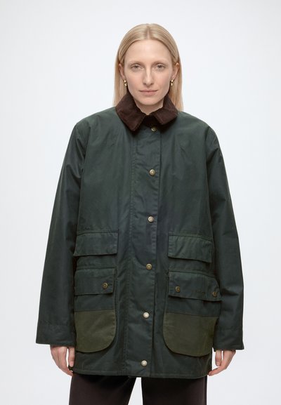 Femme aux cheveux blonds raides porte une veste vert foncé avec un col en velours côtelé marron et de grandes poches frontales, debout devant un fond uni.