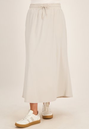 Persoon draagt een lange, lichtbeige rok met elastische tailleband en trekkoord, gecombineerd met witte sneakers op een effen achtergrond.