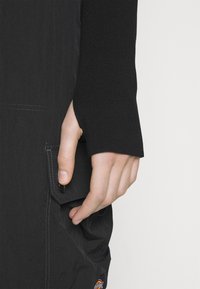 Main à la peau pâle portant un vêtement à manches longues noir, reposant sur un pantalon noir avec une poche à rabat et le logo Dickies en bas.