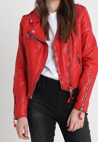 Persoon in een felrode leren jas boven een witte shirt en zwarte spijkerbroek, met ontspannen handen langs de zij.