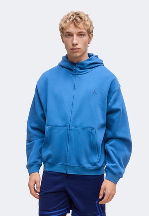 Blonder junger Mann trägt einen blauen Kapuzenpullover mit Reißverschluss und dunkelblaue Sportshorts, steht vor einem schlichten Hintergrund.