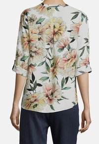 Blusa bianca con maniche arrotolate, decorata con grandi pattern floreali nei toni del giallo pastello e del rosa, accompagnati da foglie verdi, mostrata da dietro su una persona.