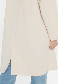 Beiger langer Mantel aus strukturiertem Stoff, mit geradem Schnitt und Seitenschlitzen, kombiniert mit hellblauer Jeans. Einfaches Design und neutrale Farbe.