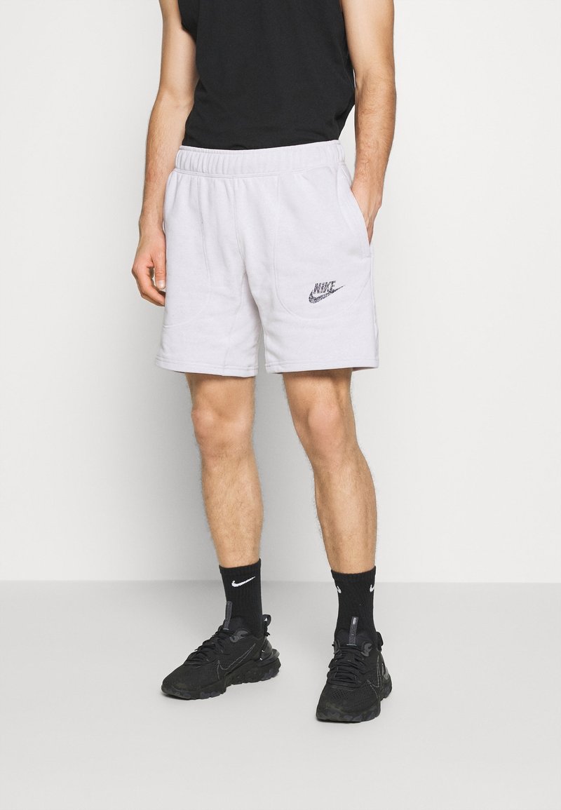 Homme portant un short Nike gris clair, des chaussettes Nike noires et des baskets noires, debout devant un fond clair uni.