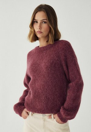 Vrouw met schouderlang haar die een zachte, lange mouwen pruimen trui en lichtbeige broek draagt, staande tegen een effen achtergrond.