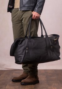 Black leather duffel bag with a textured finish, gold hardware accents, dual top handles, and two front zip pockets, held by a person.