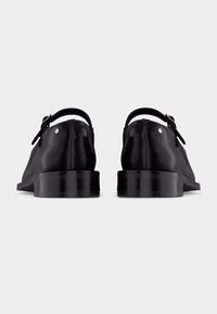 Μαύρα δερμάτινα παπούτσια φόρμας με τακούνια, ρυθμιζόμενους ιμάντες αστραγάλου και λεπτομέρειες με ασημένιες αγκράφες, εμφανιζόμενα από πίσω σε λευκό φόντο.