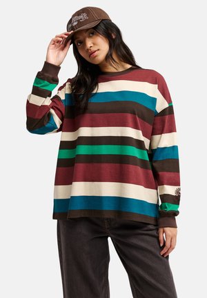 Junge Frau trägt ein mehrfarbig gestreiftes Langarmshirt, dunkle Hose und eine braune Kappe, richtet die Kappe mit einer Hand.