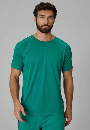 Hombre con cabello oscuro y rizado y barba, que lleva una camisa verde lisa de manga corta y pantalones verdes a juego sobre un fondo gris.
