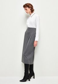Grauer Bleistiftrock mit Falten, kombiniert mit einem weißen Hemd mit Kragen. Modell trägt schwarze kniehohe Stiefel mit Absatz. Weiche Textur, taillierte Passform.