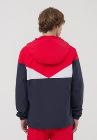 Giacca con cappuccio rossa, bianca e blu navy con un design a zigzag sul retro. Presenta polsini elastici e un fit casual. Materiale morbido e leggero.