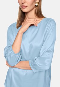 Light blue satin blouse with a round neckline and three-quarter elastic sleeves. Smooth texture with a relaxed fit and minimal detailing.