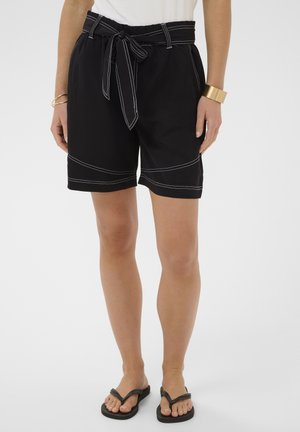 Kvinde iført sorte knælange shorts med hvide syninger, bindebælte i taljen, hvid top, guldbangle og sorte klipklapper på hvid baggrund.