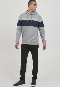 Grå hettegenser med horisontale blå og marineblå striper, lange ermer, justerbar snor, kombinert med svarte bukser og svarte sneakers.